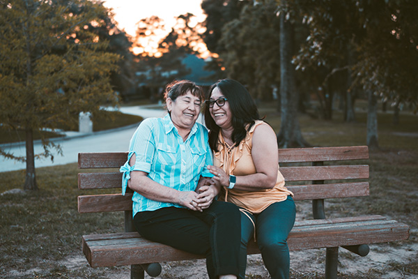 A woman sits with her older mother on a park bench.