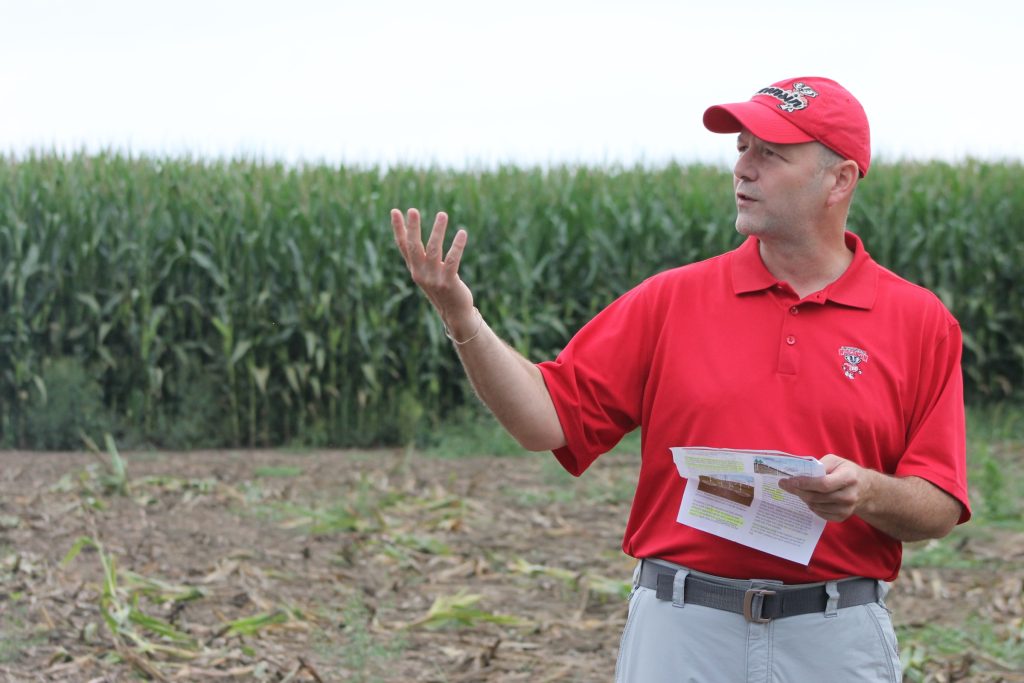 Person in a red shirt and red hat standing in a corn field giving a presentation