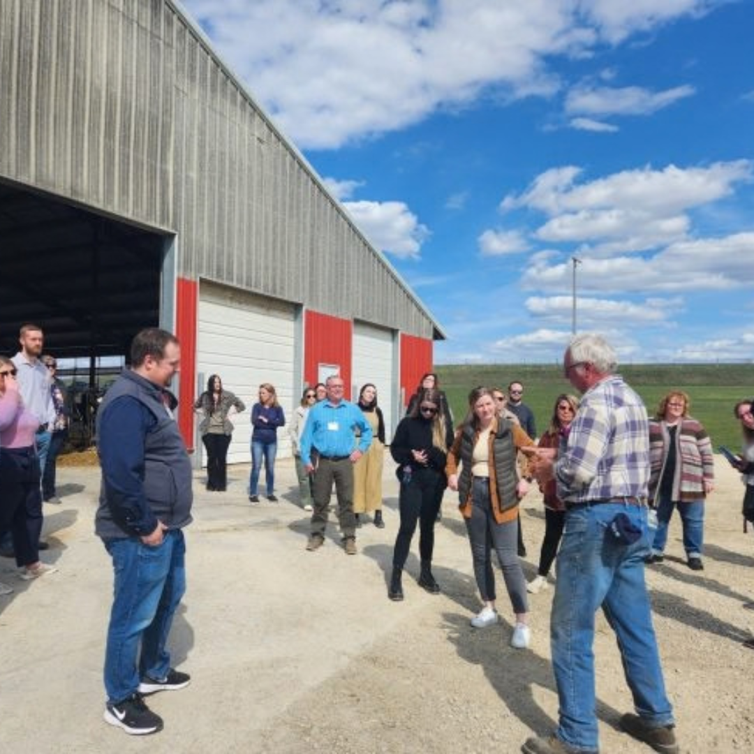 Group of people listening to a man speaking while standing by a farm building