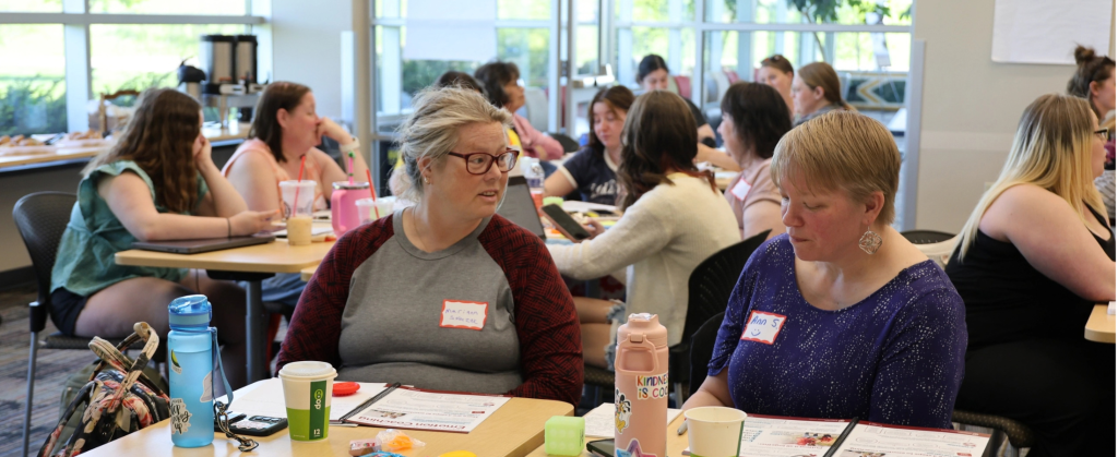 Two women sit at a table in a room full of people at tables and discuss