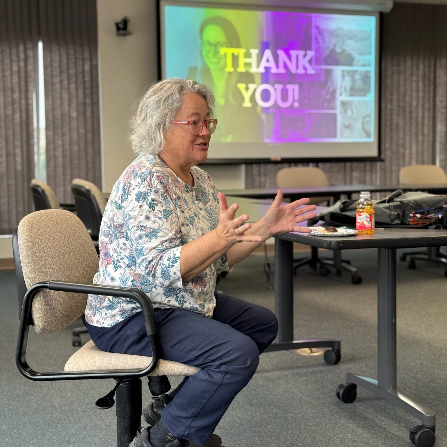 Woman sitting in a chair and talking with a message on a screen behind her that says "Thank you"