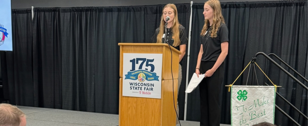 Two people standing at a podium on a stage. One person is speaking into the microphone